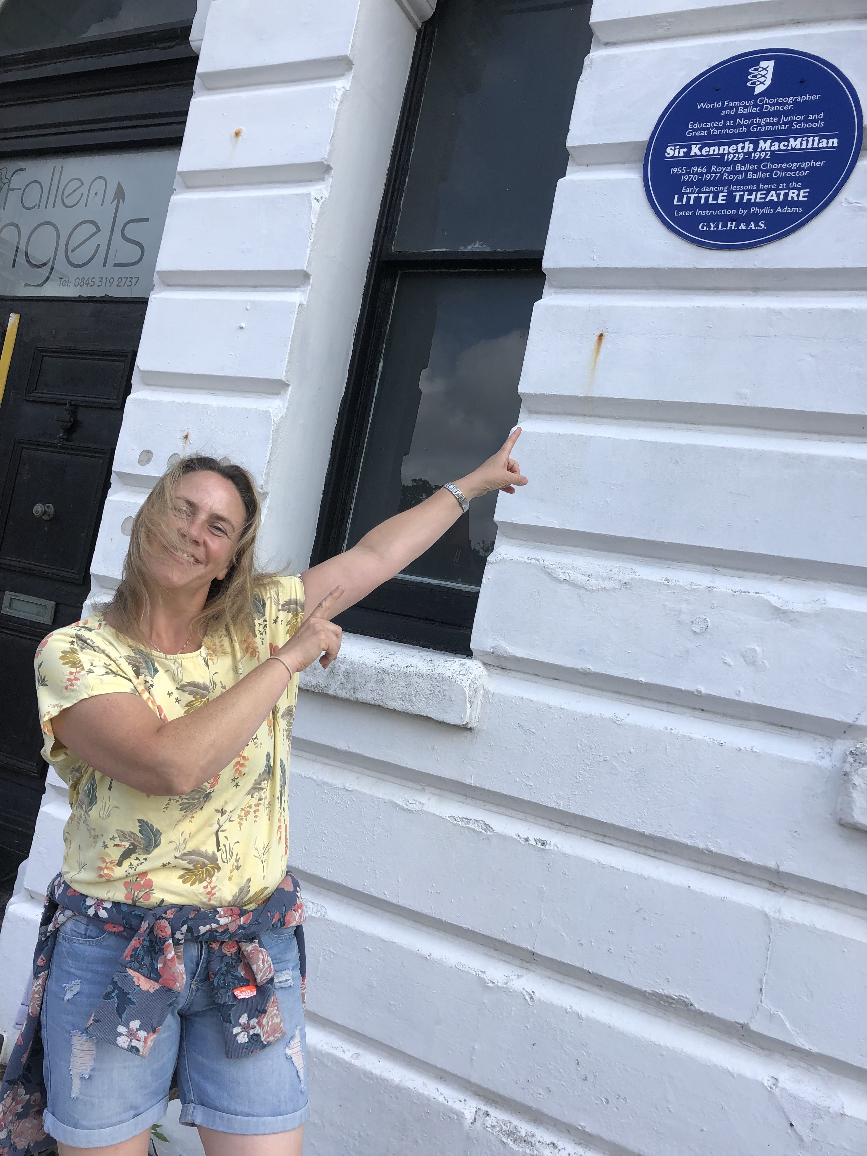 Woman standing in front of and pointing towards a Blue Plaque.